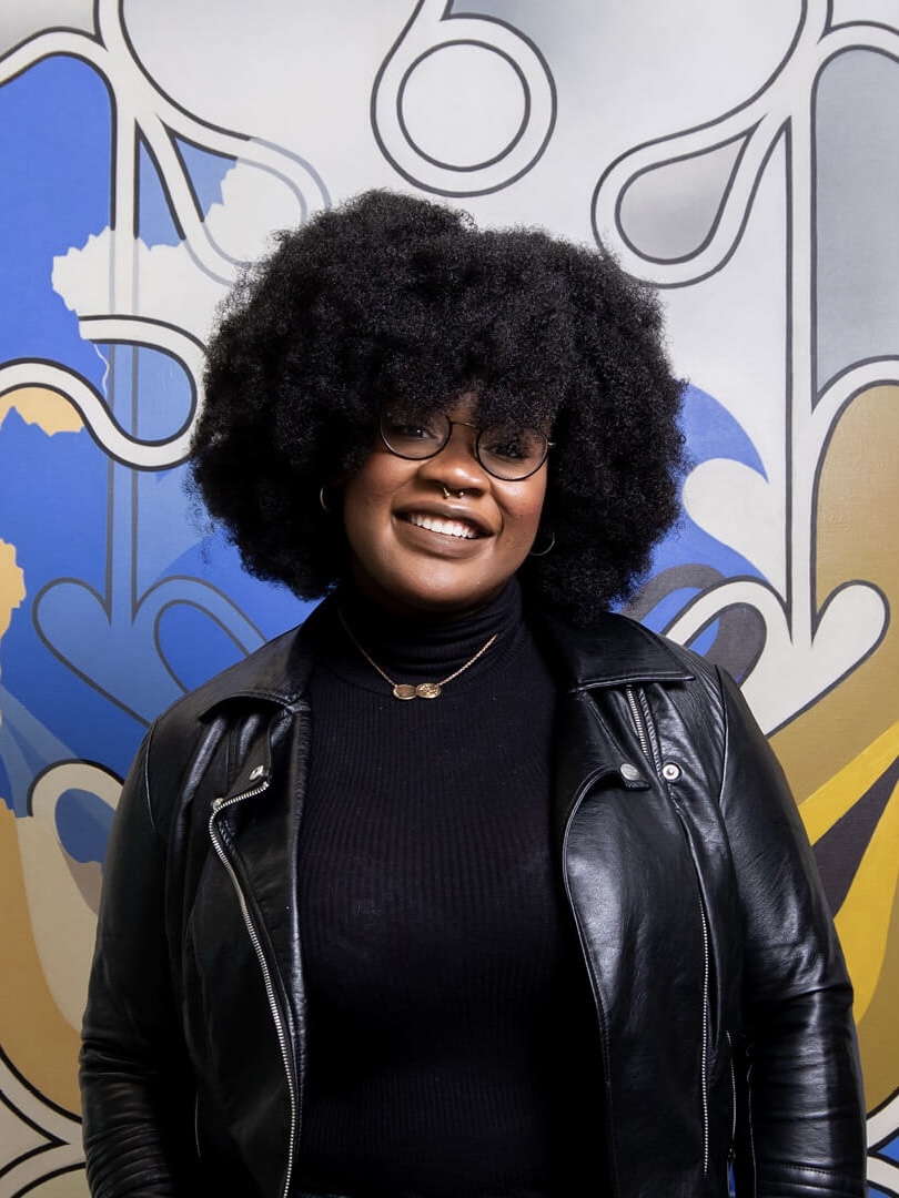 woman with round glasses against a colorful background with a black turtleneck and black leather jacket