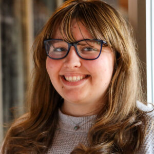 woman with light brown hair and bangs and glasses, wearing a grey sweater.