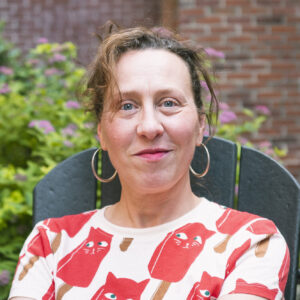 Woman sitting outside in a chair with a brick wall and bushes behind her