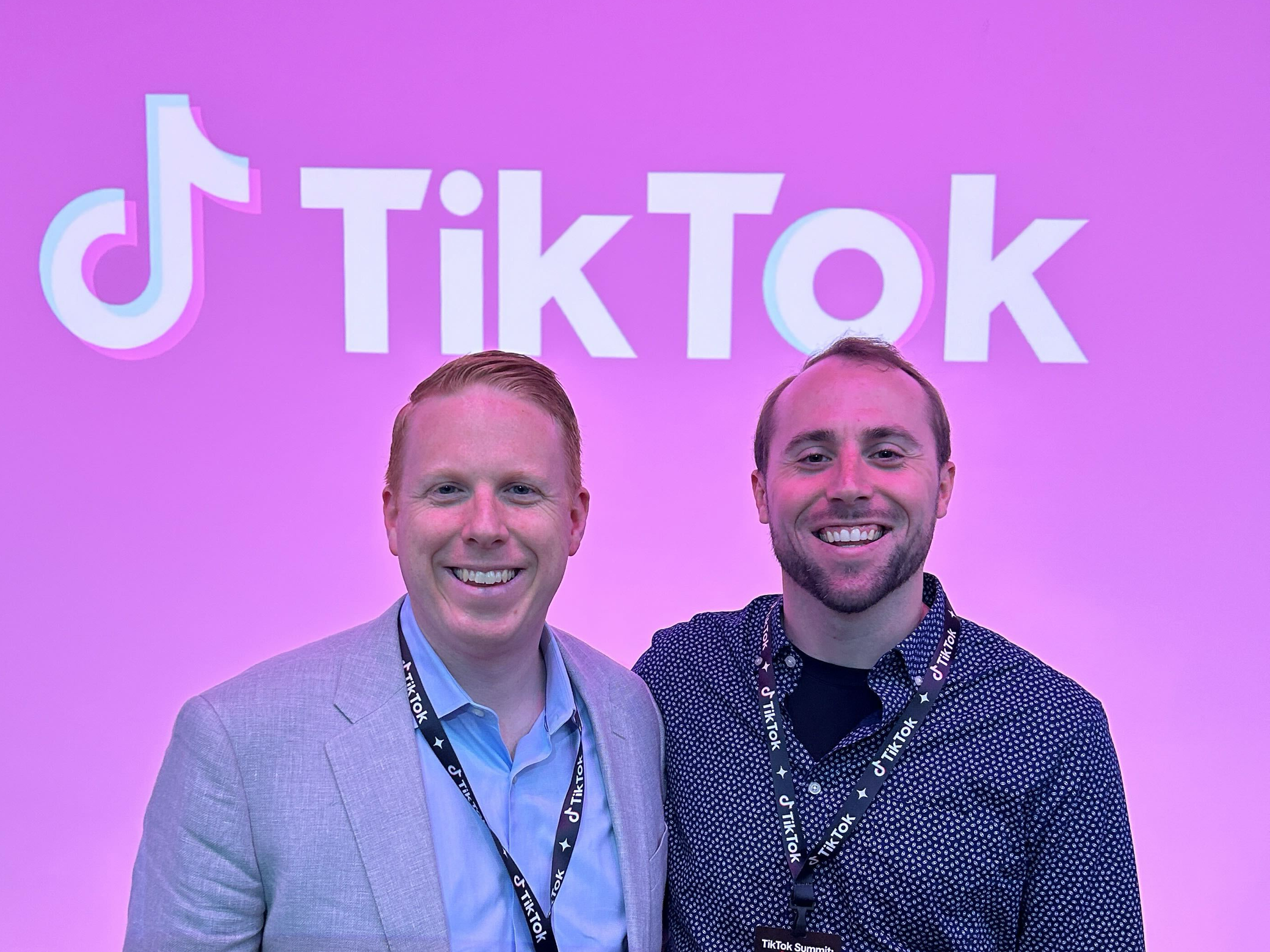 two men standing in front of a pink backdrop with TikTok in the background