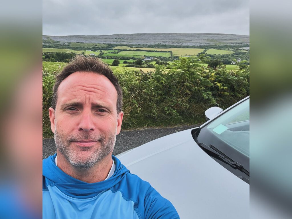 man standing on side of road with green fields behind